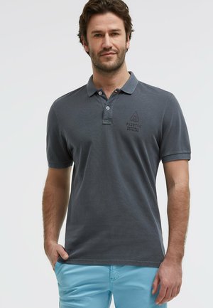 Mann trägt dunkelgraues Kurzarm-Poloshirt mit Logo auf der Brust und hellblaue Hose, steht mit einer Hand in der Tasche.
