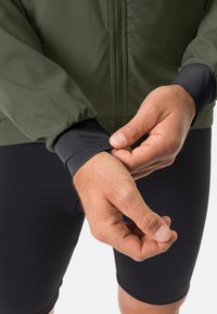Grön lättviktsjacka med svarta muddar, med dragkedja framtill. Materialet ser jämnt ut med en subtil textur. Svarta åtsittande shorts nedanför.