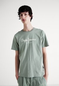 Joven vestido con una camiseta verde claro de Pepe Jeans London y pantalones a juego, de pie frente a un fondo blanco liso.
