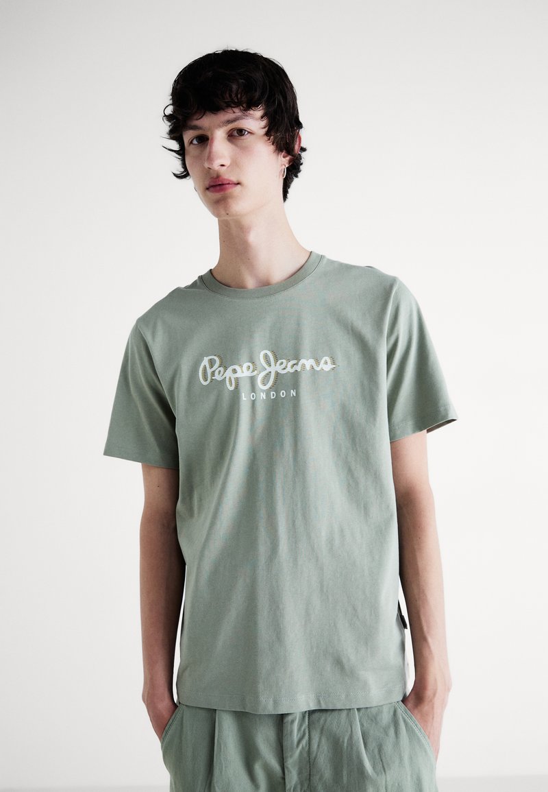Joven vestido con una camiseta verde claro de Pepe Jeans London y pantalones a juego, de pie frente a un fondo blanco liso.