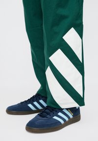 Πράσινο παντελόνι με μεγάλες λευκές διαγώνιες ρίγες στα κάτω πόδια, συνδυασμένο με σκουρόχρωμα μπλε αθλητικά παπούτσια που έχουν ανοιχτό μπλε ρίγες και χρυσή λεπτομέρεια.