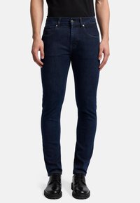 Jeans in denim blu scuro con vestibilità slim, dotati di cinque tasche, bottone in metallo e rivetti, e una leggera texture sbiadita. Abbinati a scarpe nere.