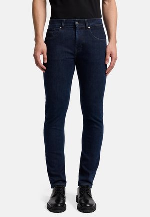 Jeans in denim blu scuro con vestibilità slim, dotati di cinque tasche, bottone in metallo e rivetti, e una leggera texture sbiadita. Abbinati a scarpe nere.