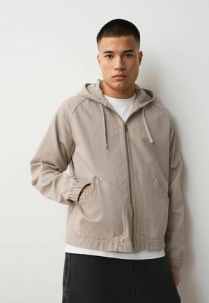 Junger Mann lehnt an einer weißen Wand, trägt eine beige Kapuzenjacke mit Reißverschluss, ein weißes Hemd und eine dunkle Hose, mit einer Hand in der Jackentasche.