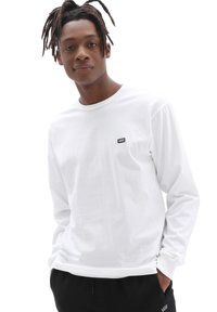 Långärmad vit T-shirt i bomull. Har en liten svart Vans-logotyp i lappen på vänster bröst. Bärs med svarta byxor.