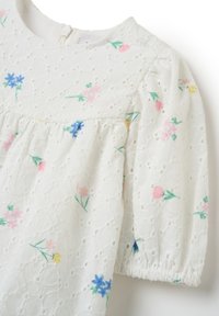 Blusa bianca con ricami floreali colorati, tessuto texturizzato con design a occhiello, maniche a sbuffo corte e chiusura con zip sulla schiena.