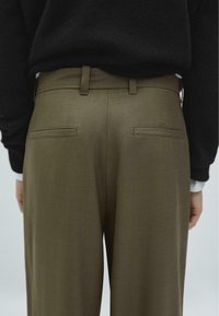 Olive green trousers with a smooth texture, featuring two back pockets with straight edges and a slim waistband. Worn with a black sweater.