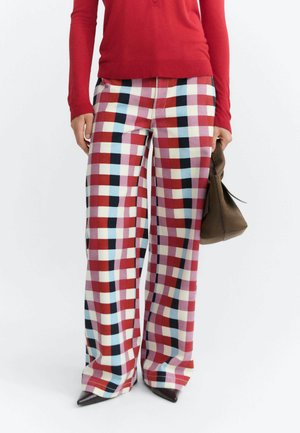 Persona che indossa una maglietta rossa a maniche lunghe e pantaloni larghi a quadri multicolori, tenendo una borsa marrone con scarpe nere con punta.