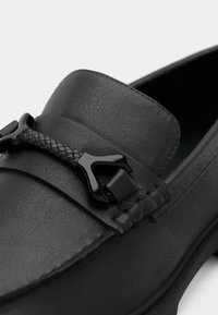 Μαύρα δερμάτινα μοκασίνια με υφή, που διαθέτουν πλεκτή διακόσμηση και γυαλιστερά μαύρα μεταλλικά στοιχεία. Στρογγυλεμένο σχήμα και λεπτομέρειες με ραφές.