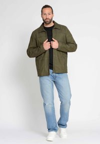 Olivengrüne Wildlederjacke mit einem Reißverschluss vorne, zwei seitlichen Taschen und einem spitzen Kragen, getragen über einem schwarzen Shirt, kombiniert mit hellblauen Jeans und weißen Sneakers.