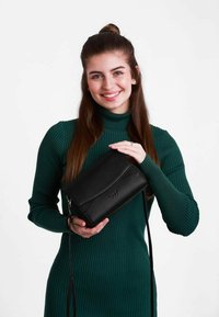 Černá kožená crossbody taška s hladkým povrchem, s klopou a embosovaným logem, držená osobou v zelených žebrovaných šatech.