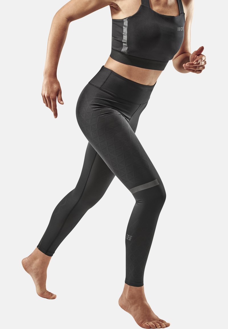Ensemble de vêtements de sport noirs, comprenant un crop top ajusté et un legging avec une texture géométrique et des accents réfléchissants. Pose pieds nus en mouvement.