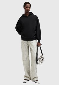 Sudadera negra con capucha de corte relajado, combinada con unos pantalones vaqueros de pierna ancha en gris claro y zapatos plateados. Sosteniendo un bolso metálico con una correa negra.