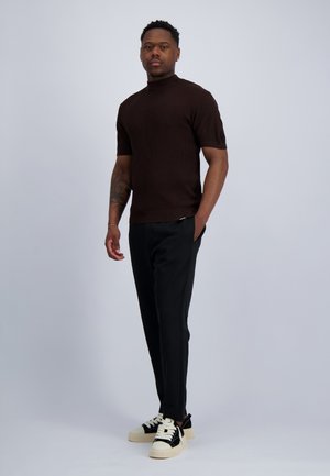 Man staat, draagt een bruin geribd shirt met korte mouwen, zwarte broek en zwart-witte sneakers, één hand in de zak.
