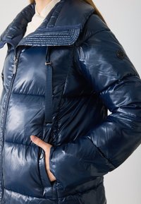 Dunkelblauer Pufferjacke mit glänzender Textur, hohem Kragen, Reißverschluss vorne und elastischen Details. Enthält Seitentaschen und ein Logo auf dem Ärmel.