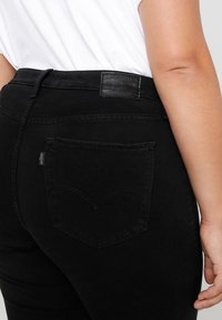 Personne portant un jean noir taille haute et une chemise blanche, montrant la poche arrière et l'étiquette en cuir Levi's sur la ceinture.