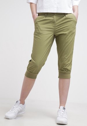 Person wearing olive green cropped pants with cuffed hems and white Adidas sneakers, standing with hands in pockets.