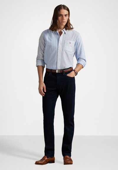 Man met schouderlang haar in een blauw overhemd met gemengde patronen, donkere jeans, een bruine riem, een horloge en bruine leren instappers.