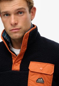 Fleece-Pullover mit marinefarbigem Körper und orangefarbenem Kragen. Verfügt über eine knöpfbare Brusttasche und ein kariertes Innenfutter. Texturierter Stoff.