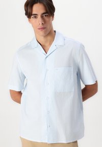 Lichtblauw kortemouwenshirt met een knoopsluiting aan de voorkant, verticale strepen en een patchzakje op de linkerborst. Zachte stof structuur.
