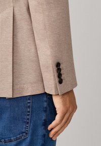 Hellbeiger Blazer mit strukturiertem Finish, ausgestattet mit schwarzen Knopf-Akzenten an den Manschetten und einem schlichten, taillierten Design.