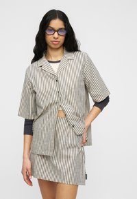 Γυναίκα με μακριά σκούρα μαλλιά που φοράει γυαλιά με μπλε απόχρωση, πουκάμισο κοντών μανικιών μπεζ και καφέ καρό και φούστα που ταιριάζει, ποζάρει με το χέρι στη μέση.