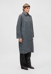 Garšs houndstooth raksta mētelis pelēkā un melnā vilnas maisījumā, ar plašu apkakli un taisnu piegriezumu, apvienots ar melnām ādas biksēm un norādītajiem apaviem.