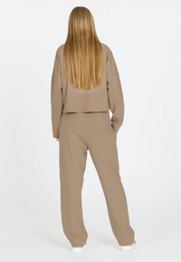 Beige cropped sweatshirt en bijpassende broek, met een ontspannen pasvorm en zachte textuur. Persoon met lang haar kijkt weg, draagt witte sneakers.