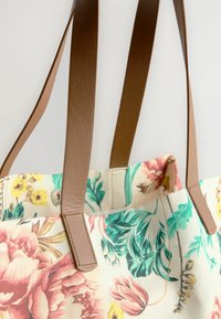 Bolsa de lona floral con asas de cuero marrón. Presenta patrones de flores y hojas multicolores sobre un fondo claro.