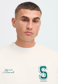 Bluza de culoare bej deschis, cu literă brodată verde, "The Ace of Diamonds," și o literă "S" texturată pe piept. Material neted.