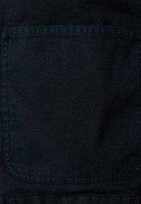 Gros plan d'une poche plaquée en tissu noir avec des coutures visibles sur un fond texturé sombre.