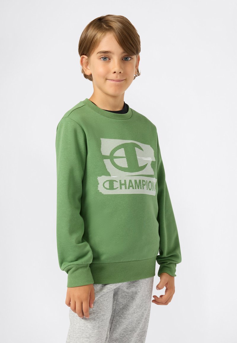 Sweatshirt vert en matériau doux, arborant un logo texturé et le mot "CHAMPION" en blanc sur la poitrine. Porté sur un pantalon de train gris.