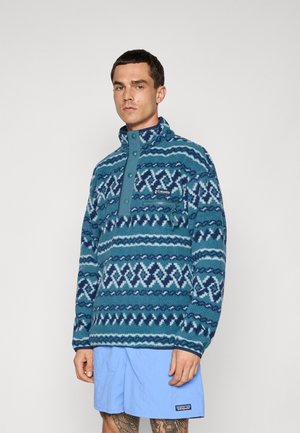 Man wearing blue patterned fleece pullover with front buttons and zip pocket, paired with light blue shorts, standing against plain background.