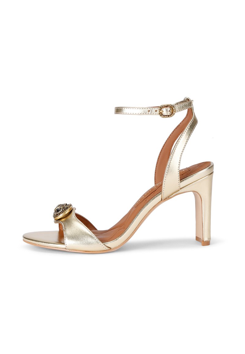 Złote metaliczne sandały na wysokim obcasie z otwartym noskiem, paskiem na kostkę i dekoracyjną okrągłą ozdobą z przodu. Gładkie wykończenie.