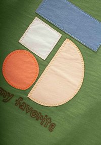 La tela verde presenta coloridos apliques geométricos: un rectángulo azul, un cuadrado blanco, un círculo naranja y un semicírculo beige, con texto marrón bordado.