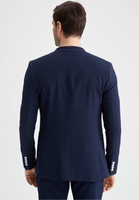 Marinblå kavaj i texturerat tyg, med enkel vent och tre knappar på varje ärm. Skräddarsydd passform som framhäver markerade axlar.