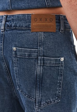 Jeans de mezclilla azul con una etiqueta de cuero OXXO. Presentan un corte clásico, bolsillos traseros visibles y acentos metálicos en los bordes de los bolsillos.