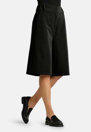 Mujer con pantalones cortos negros de pierna ancha hasta la rodilla y mocasines negros, de pie con las piernas cruzadas sobre un fondo liso.