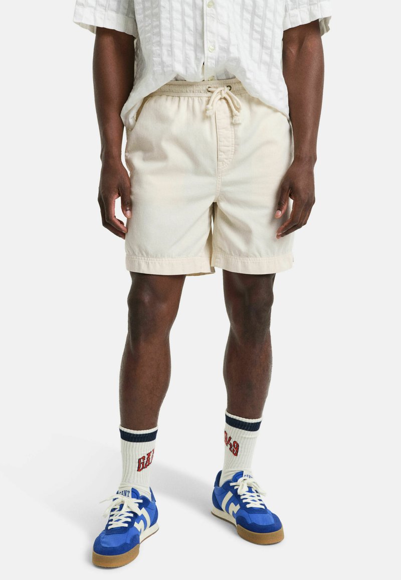 Beige bomuldsshorts med elastisk talje og snor, parret med blå sneakers og hvide sokker med mørkeblå striber og rød tekst.