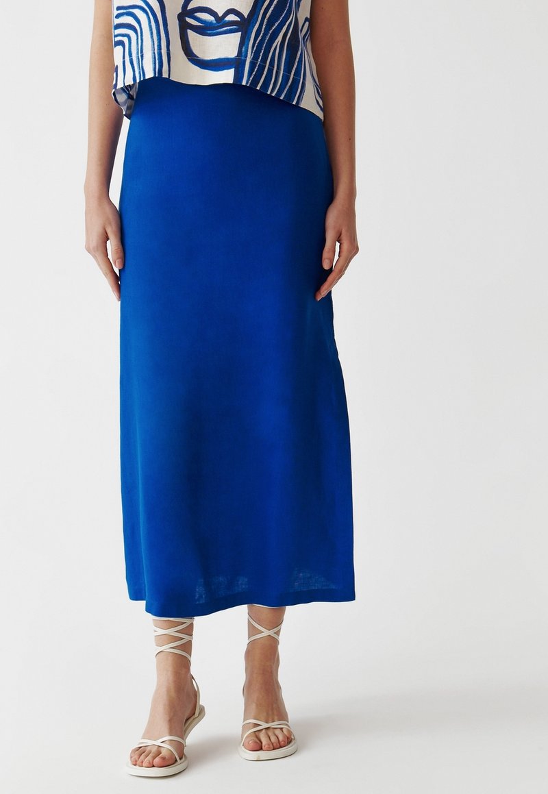Jupe longue bleue en tissu lisse, présentant une coupe trapèze simple. Portée avec des sandales blanches à lanières aux pieds.