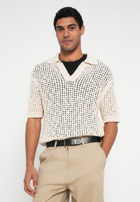 Krátký pletený svetr s krátkými rukávy v krémové barvě, s výstřihem do V a otevřeným síťovaným vzorem. Nošený přes černé tričko, v kombinaci s béžovými kalhotami a černým pásem.