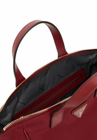 Rote Lederhandtasche mit goldfarbener Reißverschluss, ausgestattet mit einem grauen Innenfutter, das mit dem Markennamen "GUESS" bedruckt ist und einer Innentasche mit Reißverschluss.