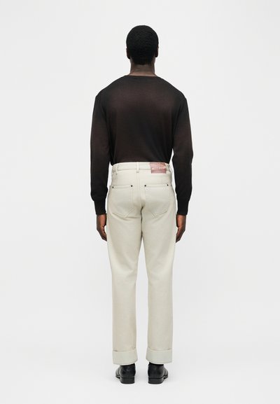 Άνδρας που φοράει μαύρο μακρυμάνικο πουκάμισο, μπεζ παντελόνι με γυρισμένα μανίκια και μαύρα παπούτσια, στέκεται αντικρίζοντας ένα απλό ανοιχτό γκρι φόντο.