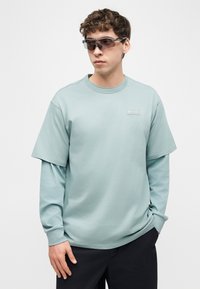 Νέος άνδρας που φοράει στρωμένα ανοιχτό μπλε μακρυμάνικα και κοντομάνικα πουκάμισα με μαύρα γυαλιά ηλίου, στέκεται με το ένα χέρι στην τσέπη μπροστά σε απλό φόντο.