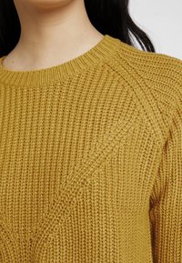 Gelber Strickpullover mit strukturiertem Muster, geripptem Kragen und lockerer Passform. Der Stoff zeigt eine leichte Dehnbarkeit und Weichheit.