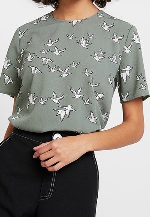 Groen shirt met korte mouwen met witte vogelzomen, gedragen in een zwarte, hooggetailleerde broek met witte stiksels en een knoopsluiting.