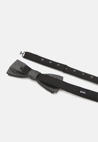 Czarny muszka z teksturowanej tkaniny z małymi, kropelek wzorami, w komplecie z regulowanym paskiem z oznaczeniami rozmiaru i etykietą z logo.