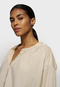 Camicia beige, leggera, a maniche lunghe con patta a bottoni, vestibilità rilassata e maniche plissettate, realizzata in tessuto morbido.