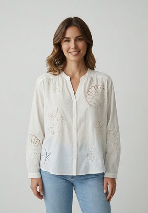 Femme souriante, portant une blouse blanche boutonnée avec une broderie de coquillage et d'étoile de mer, associée à un jean bleu clair, sur un fond uni.