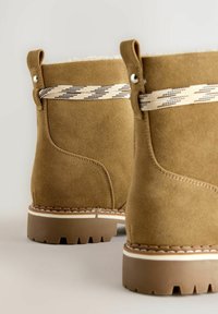 Närbild av ljusbruna mockabottnar med mönstrade beige och bruna snören, tjocka gummisulor och dragöglor bakpå.
